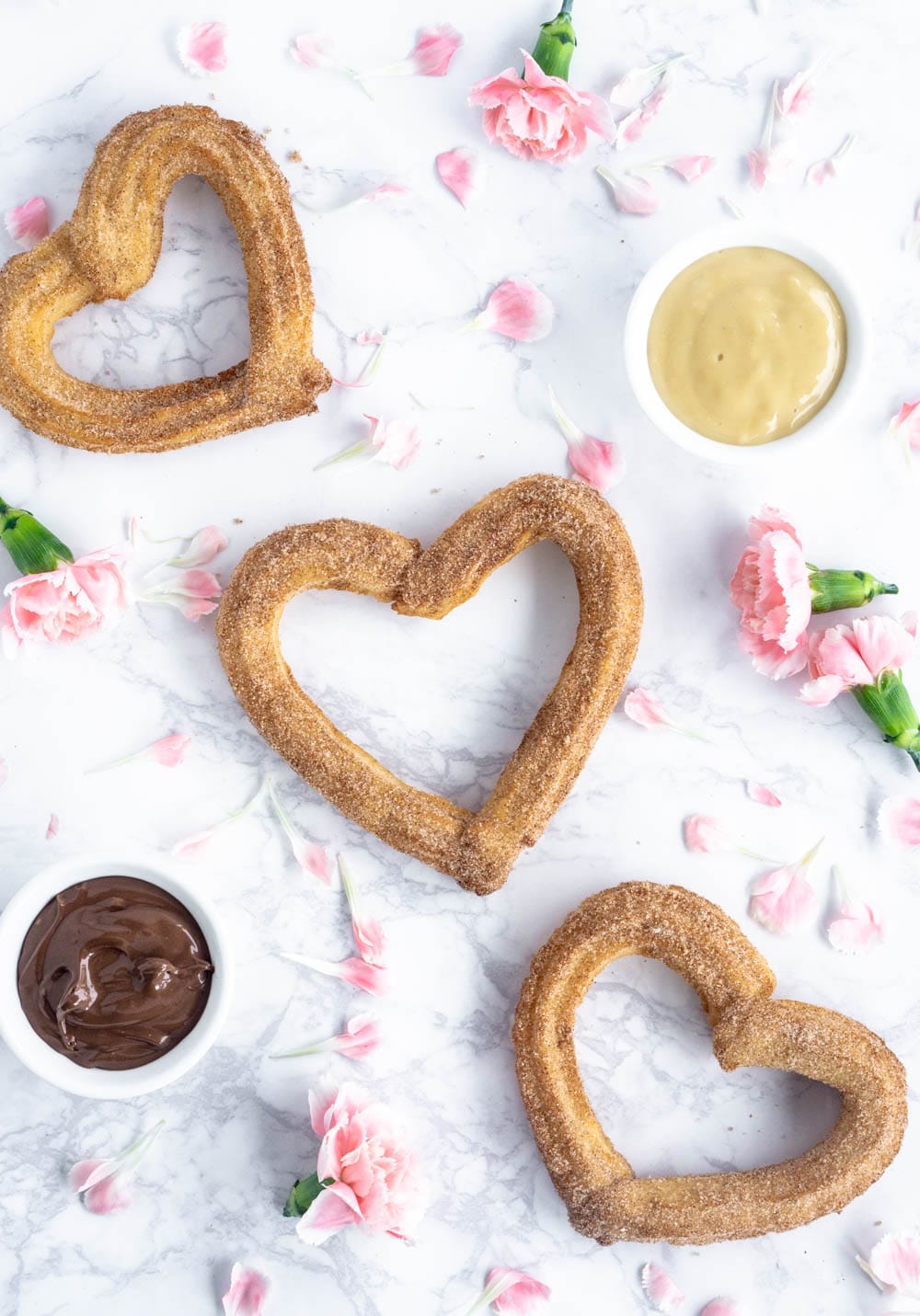Valentine's Day dessert idea: heart churros with nutella and vanilla mascarpone dipping sauces! #valentinesday #valentinesdaybaking #valentinesdaydesserts #churros #heartchurros 3 heart shaped churros on a marble background with pink flowers