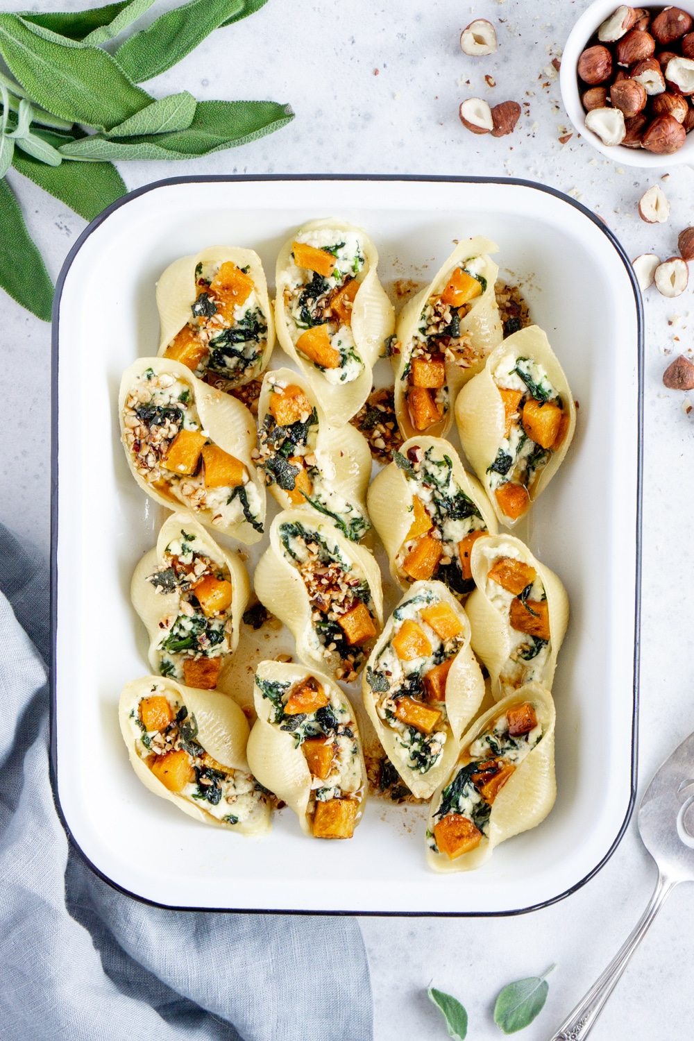 Italian stuffed pasta shells with butternut squash, spinach and ricotta. Baked and then topped with an amazing brown butter, sage and hazelnut sauce! #stuffedshells #pastashells #pinacooks Cooked stuffed pasta shells in a baking dish surrounded by fresh sage and a gray napkin