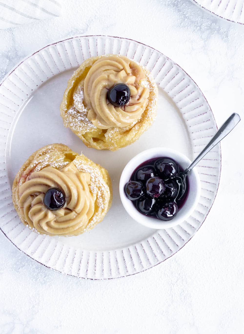 Zeppole di San Giuseppe Recipe #pinacooks #zeppoledisangiuseppe #italiandesserts overhead shot of zeppole di san Giuseppe on a white plate with amarena cherries