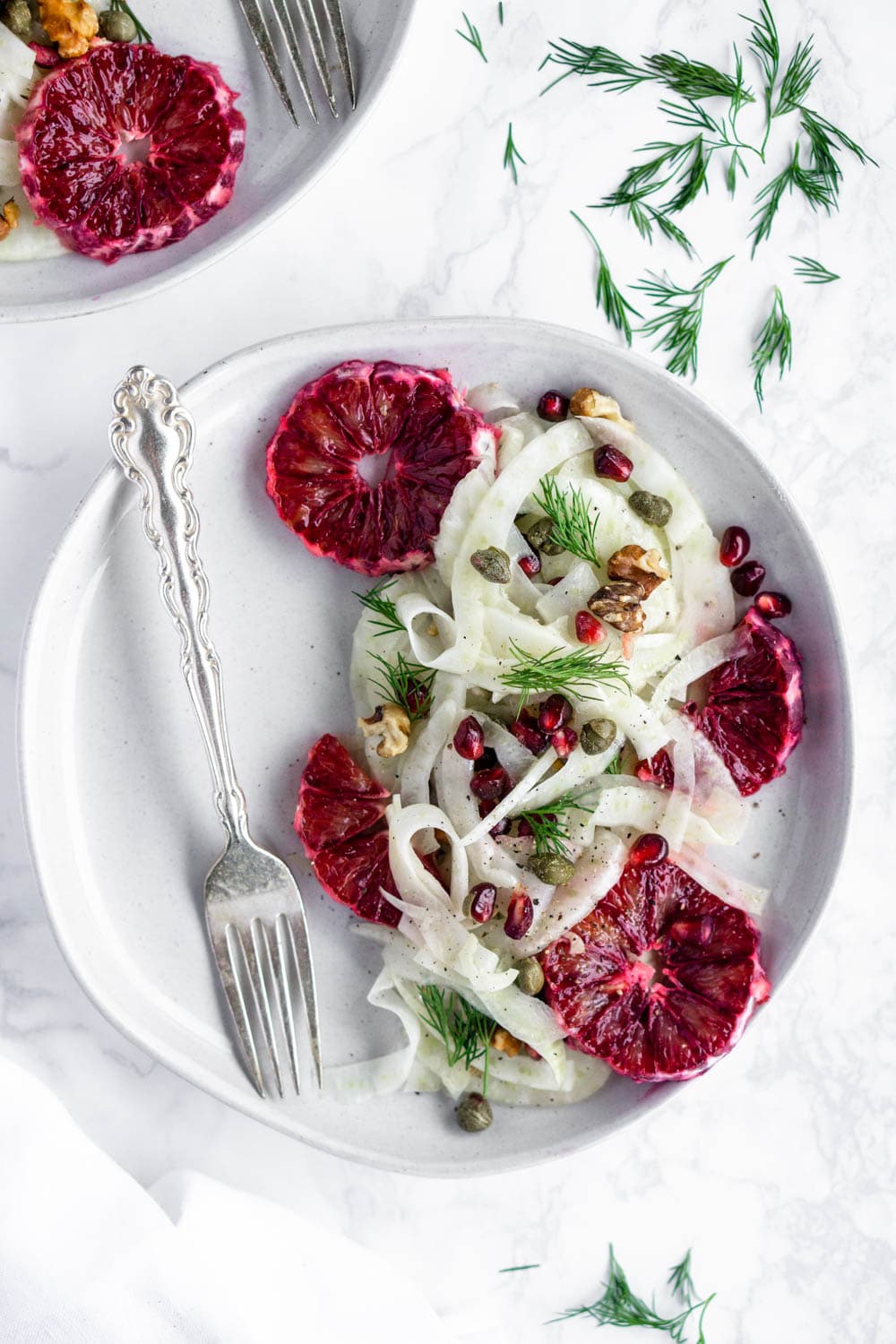 Fresh shaved fennel salad with blood orange, pomegranate, capers, walnuts and dill! So pretty too! #fennelsalad #bloodorangesalad #wintersalad #pinacooks Fennel salad on a plate with blood oranges, capers, dill and walnuts in a plate with a fork