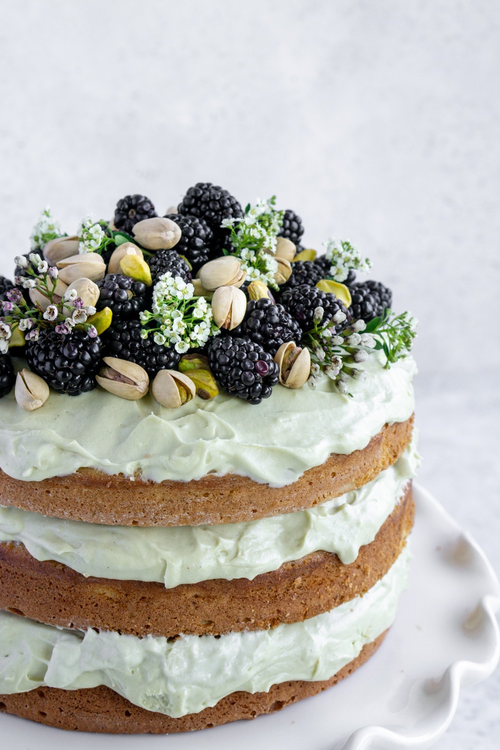 Double pistachio cake from scratch! Made with real pistachios and real ingredients. #pinacooks #pistachiocake #italiancake side view of layered pistachio cake topped with flowers, blackberries and pistachios