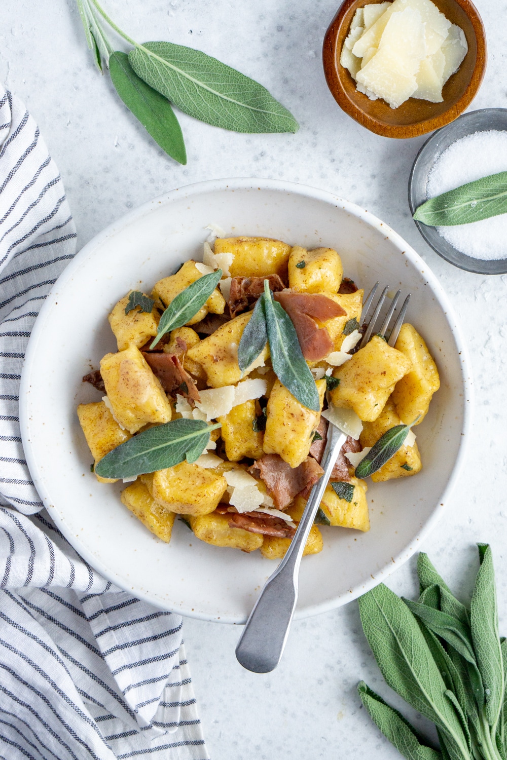 Butternut squash gnocchi recipe with brown butter, sage and prosciutto. Perfect for fall! #butternutsquashgnocchi #pinacooks #italianrecipes #fallrecipes Bowl of butternut squash gnocchi topped with sage and bowls of salt and cheese to the side