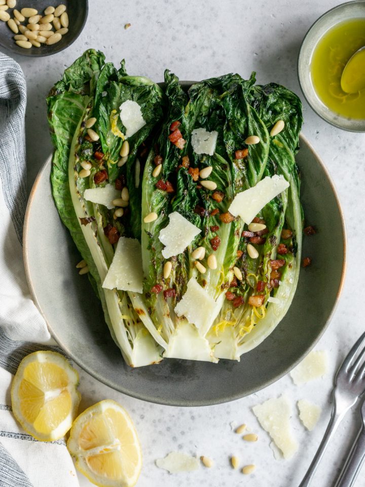 Grilled romaine salad in a bowl surrounded by lemons. pine nuts and vinaigrette