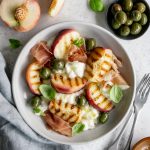 Peach burrata salad in a plate surrounded by a small plate of olives and a sliced peach