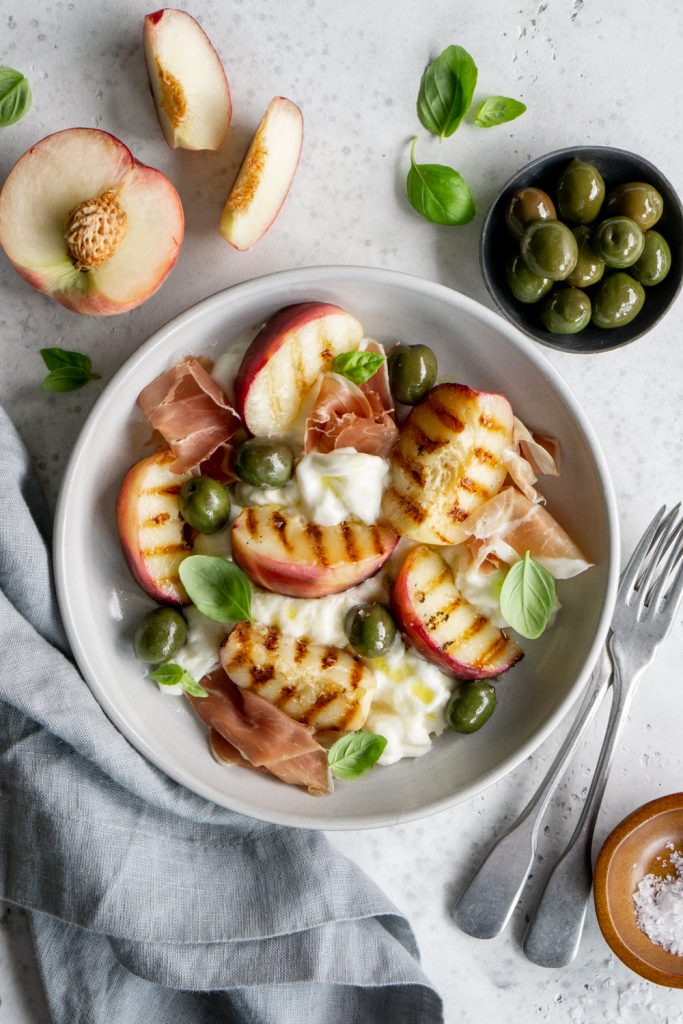 Peach burrata salad in a plate surrounded by a small plate of olives and a sliced peach
