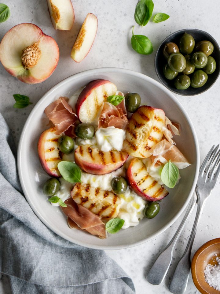 Peach burrata salad in a plate surrounded by a small plate of olives and a sliced peach