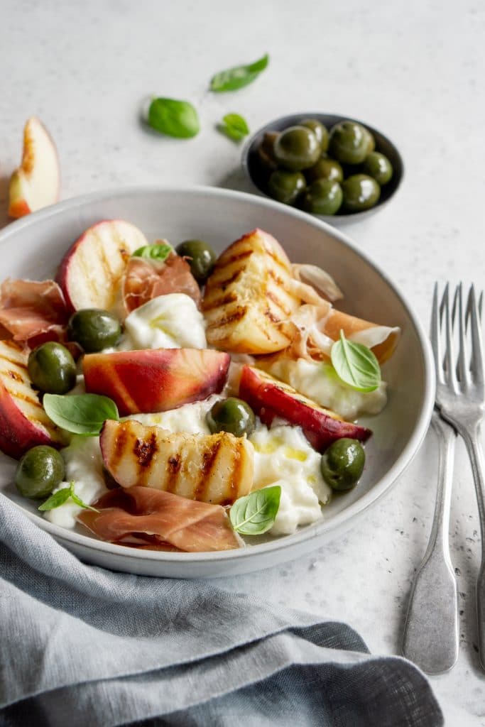 Peach burrata salad with a small plate of olives in the background and fresh basil leaves