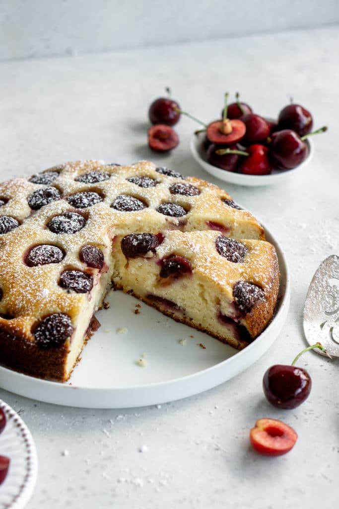 Cherry cake cut into slices on a white plate with fresh cherries in the background