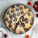Cherry cake on a white plate with 3 slices cut out of it surrounded by fresh cherries