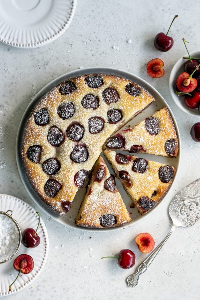 Cherry cake on a white plate with 3 slices cut out of it surrounded by fresh cherries