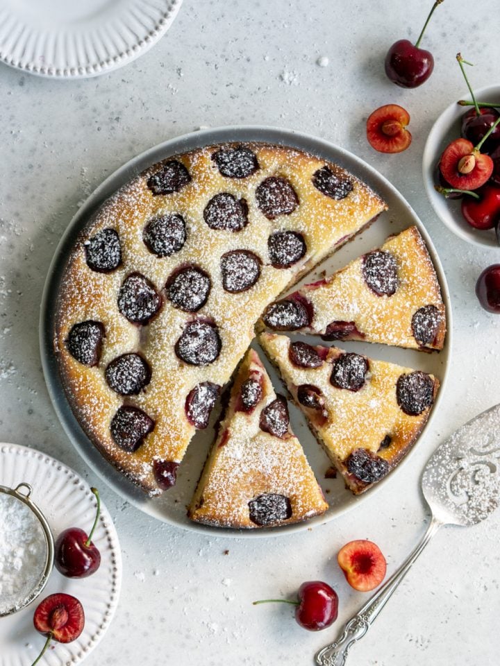 Cherry cake on a white plate with 3 slices cut out of it surrounded by fresh cherries