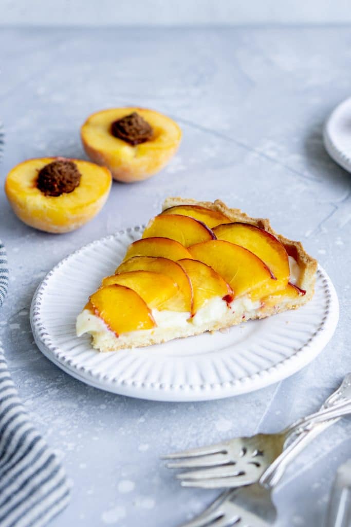 Slice of peach tart on a white plate with halved peach in the background