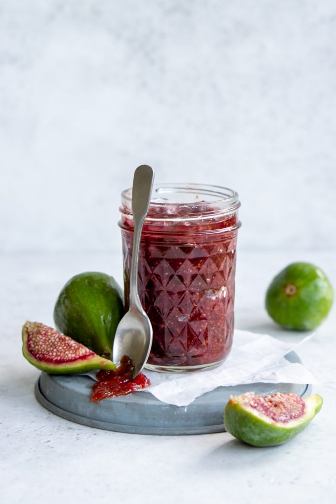 Jar of fig jam on a steel plate, surrounded by fresh figs