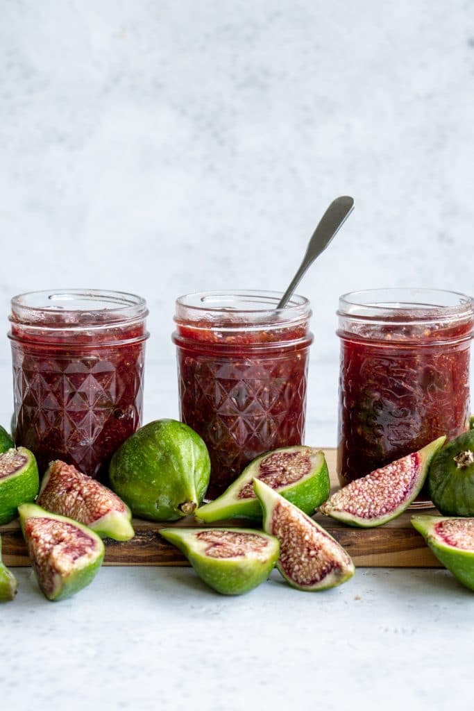 Three jars of fig jam on a wood board surrounded by fresh figs