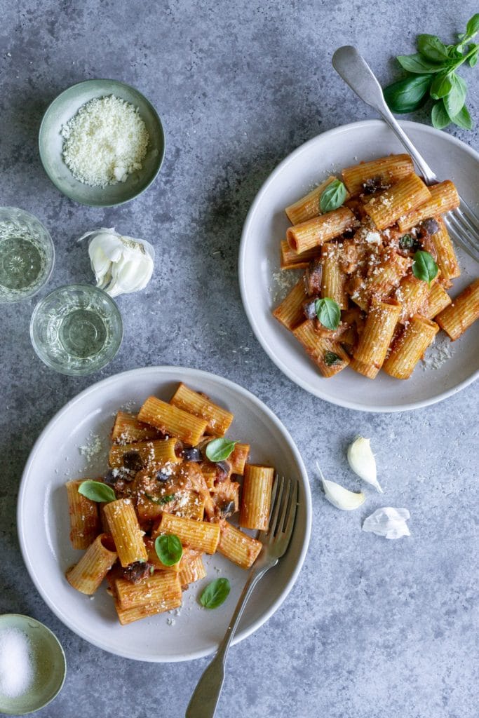 Two plates of eggplant pasta and two glasses of wine and garlic beside them