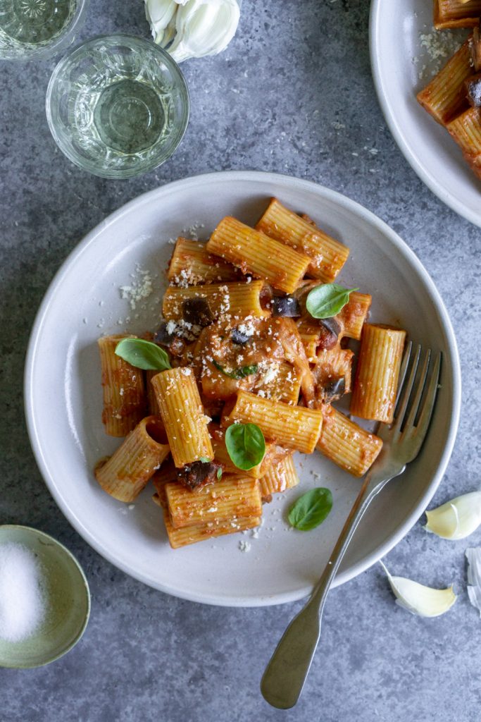 Eggplant pasta in a plate surrounded by garlic and a glass of white wine