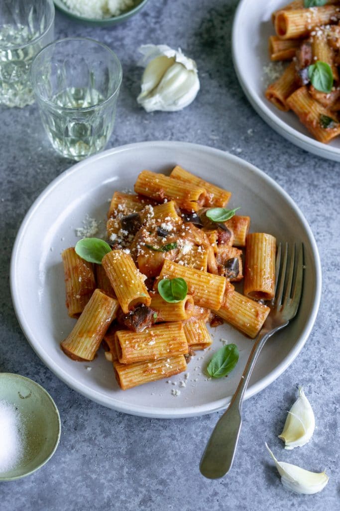 Plate of eggplant pasta topped with fresh basil and a clove of garlic in the background