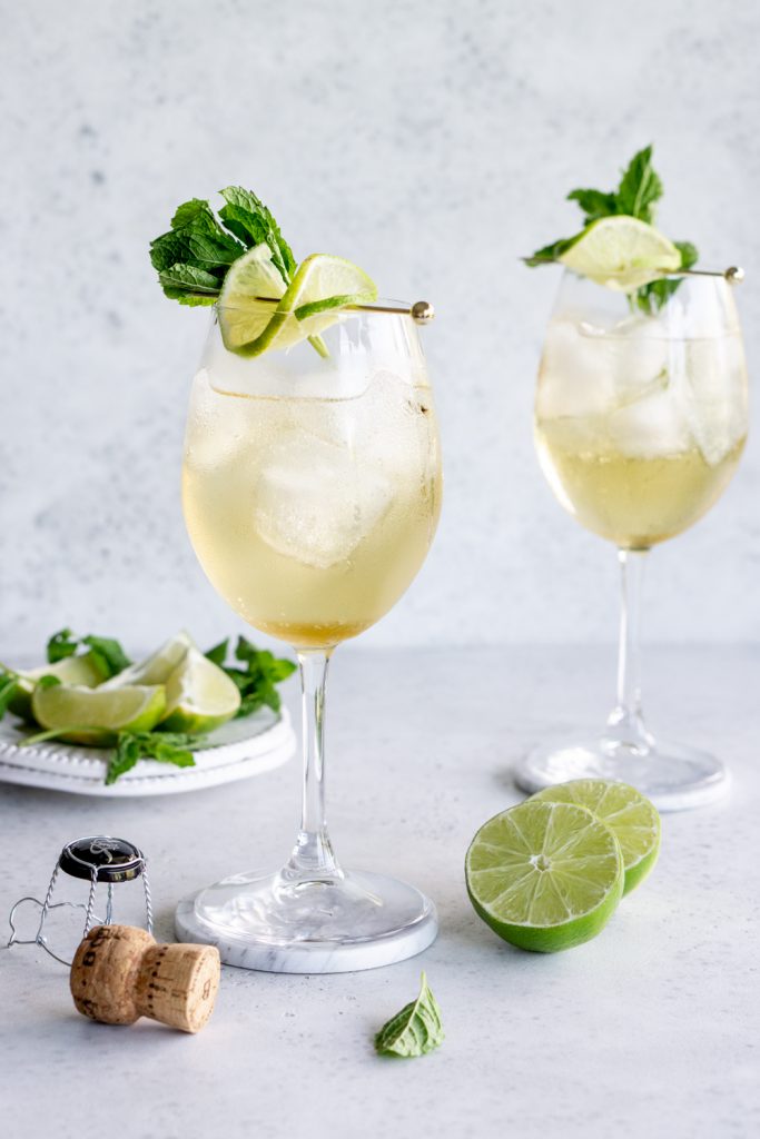 2 hugo cocktails on coasters garnished with lime and mint, with a cork to the side