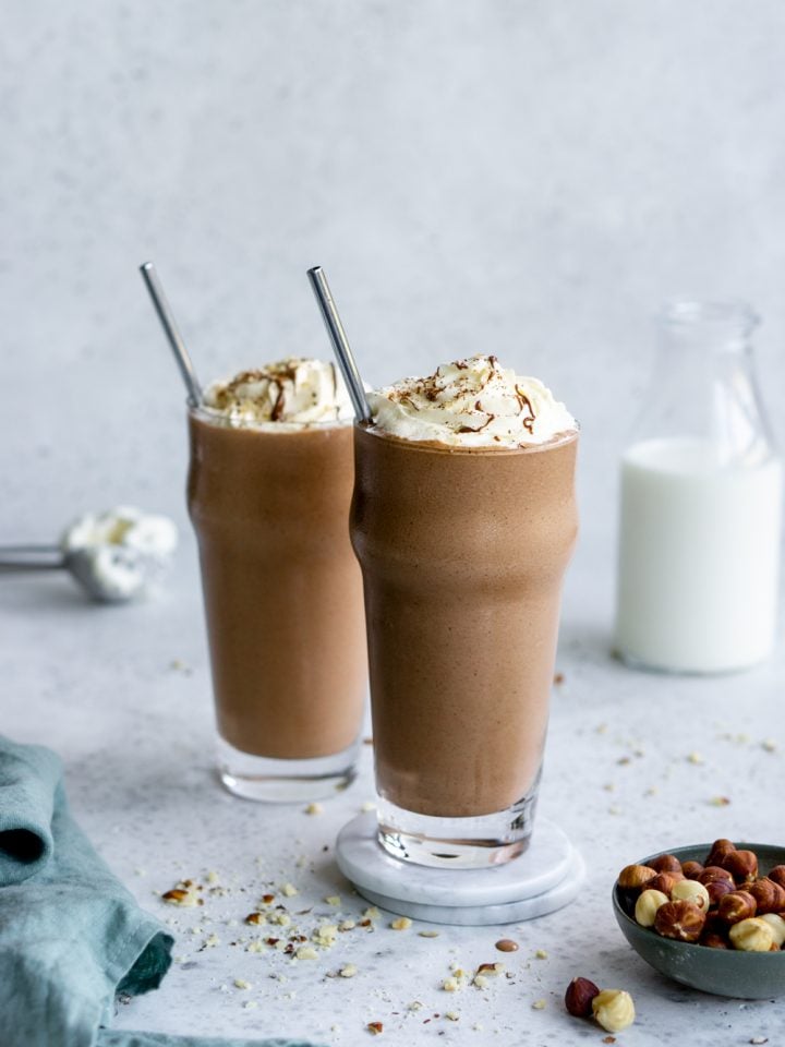 Two nutella milkshakes in tall glasses with a bowl of hazelnuts and a napkin to the side