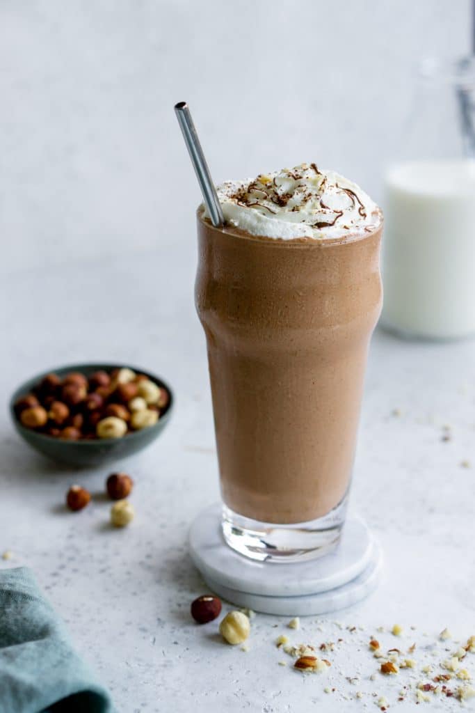 Nutella milkshake in a glass topped with whipped cream and a bowl of hazelnuts in the background