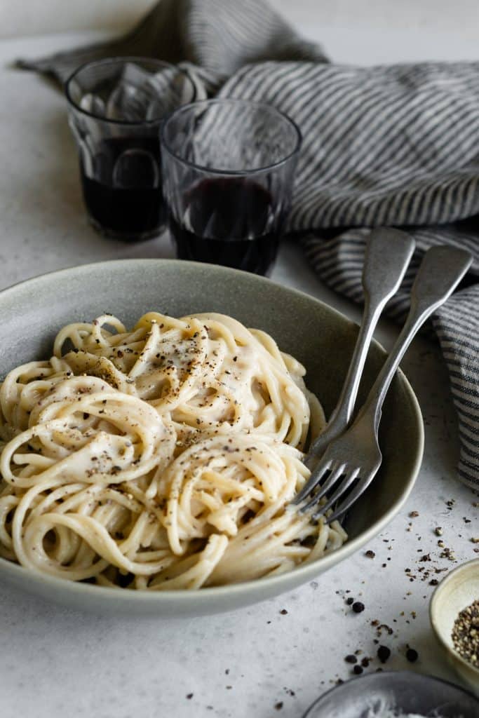 Authentic cacio e pepe in a green bowl with 2 glasses of red wine and a napkin in the background
