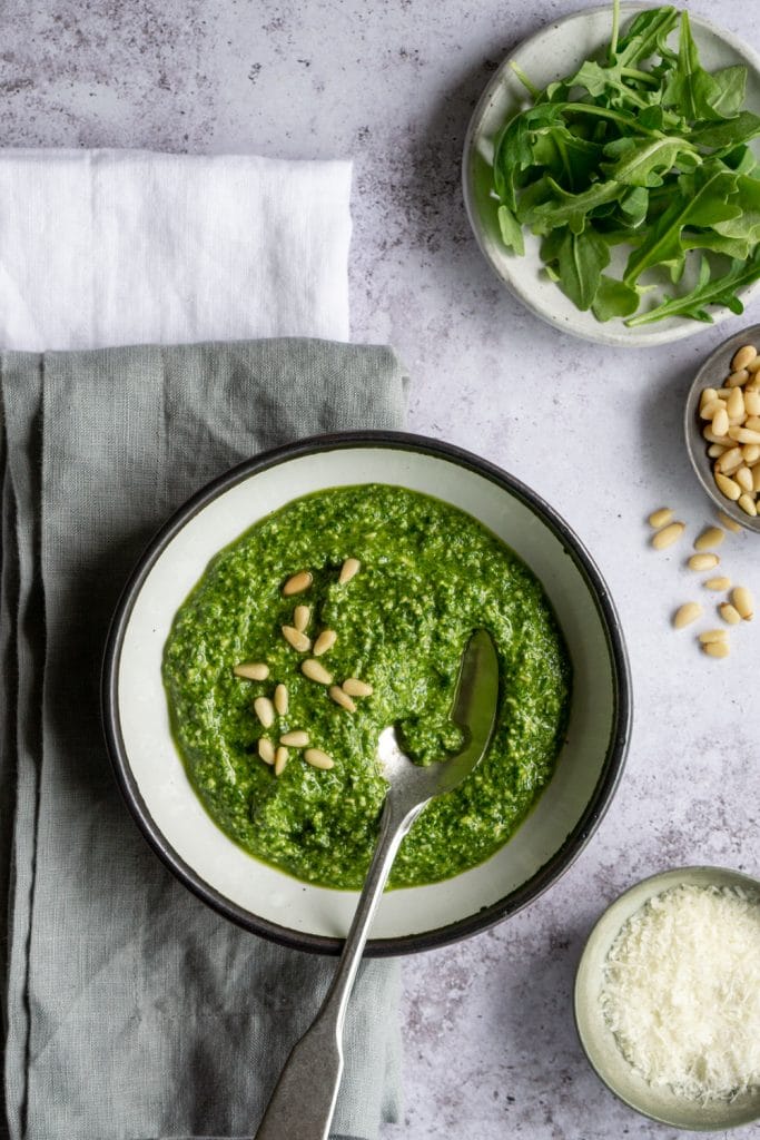 Arugula pesto in a bowl with dishes of arugula and pine nuts to the side