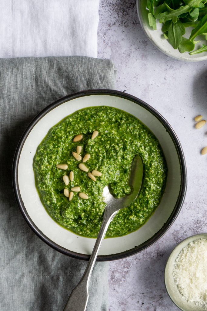 White bowl of arugula pesto topped with pine nuts, and a spoon in the bowl