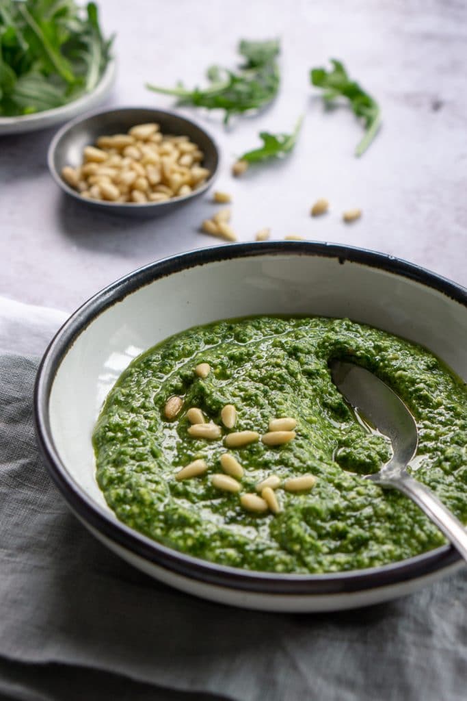 Side view of arugula pesto in a bowl with a dish of pine nuts and loose arugula in the background