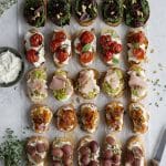 Five different types of crostini arranged on parchment paper with a knife and bowl of ricotta on either side