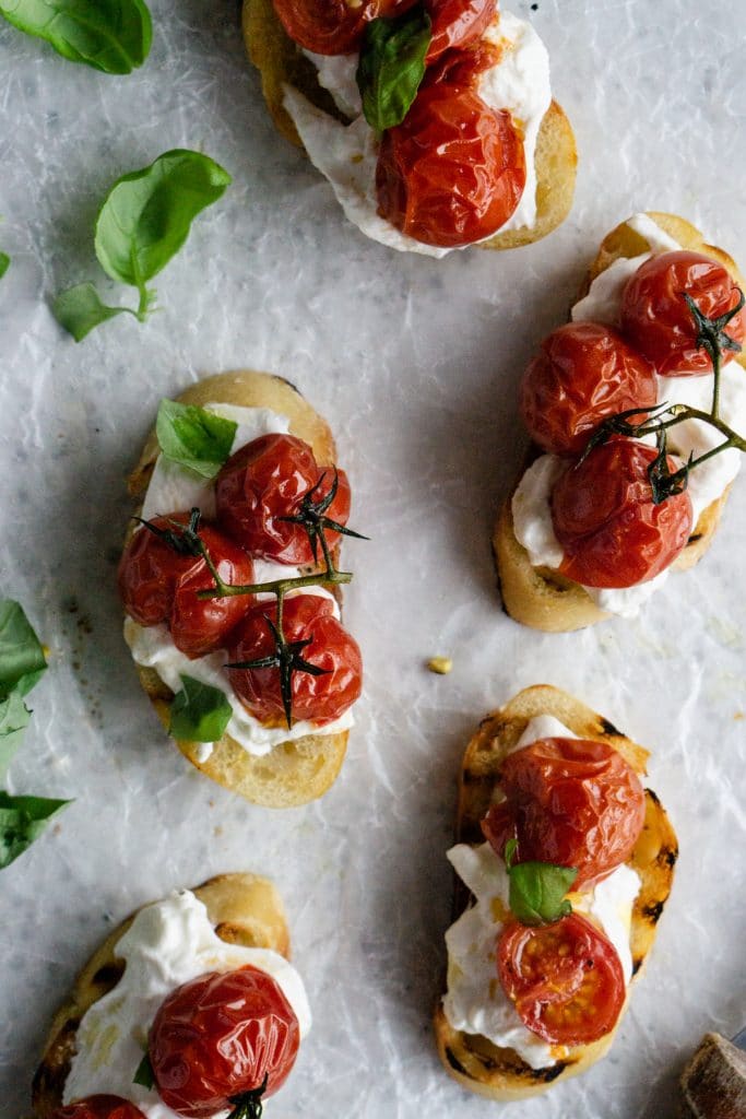 Five roasted tomato crostini on parchment paper with fresh basil to the side