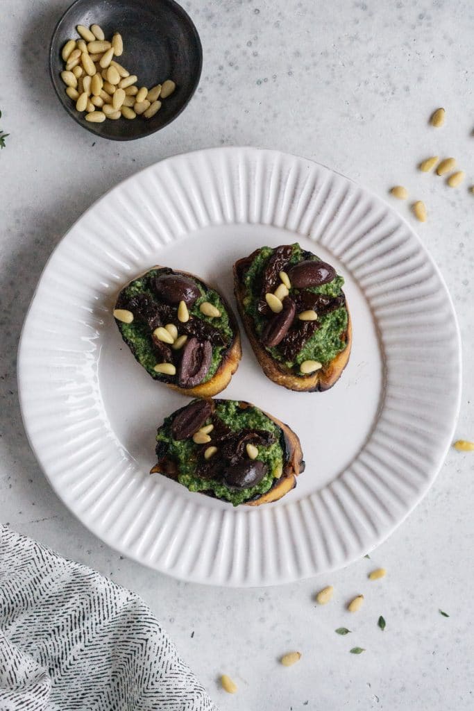 Pesto crostini on a white plate with a small bowl of pine nuts
