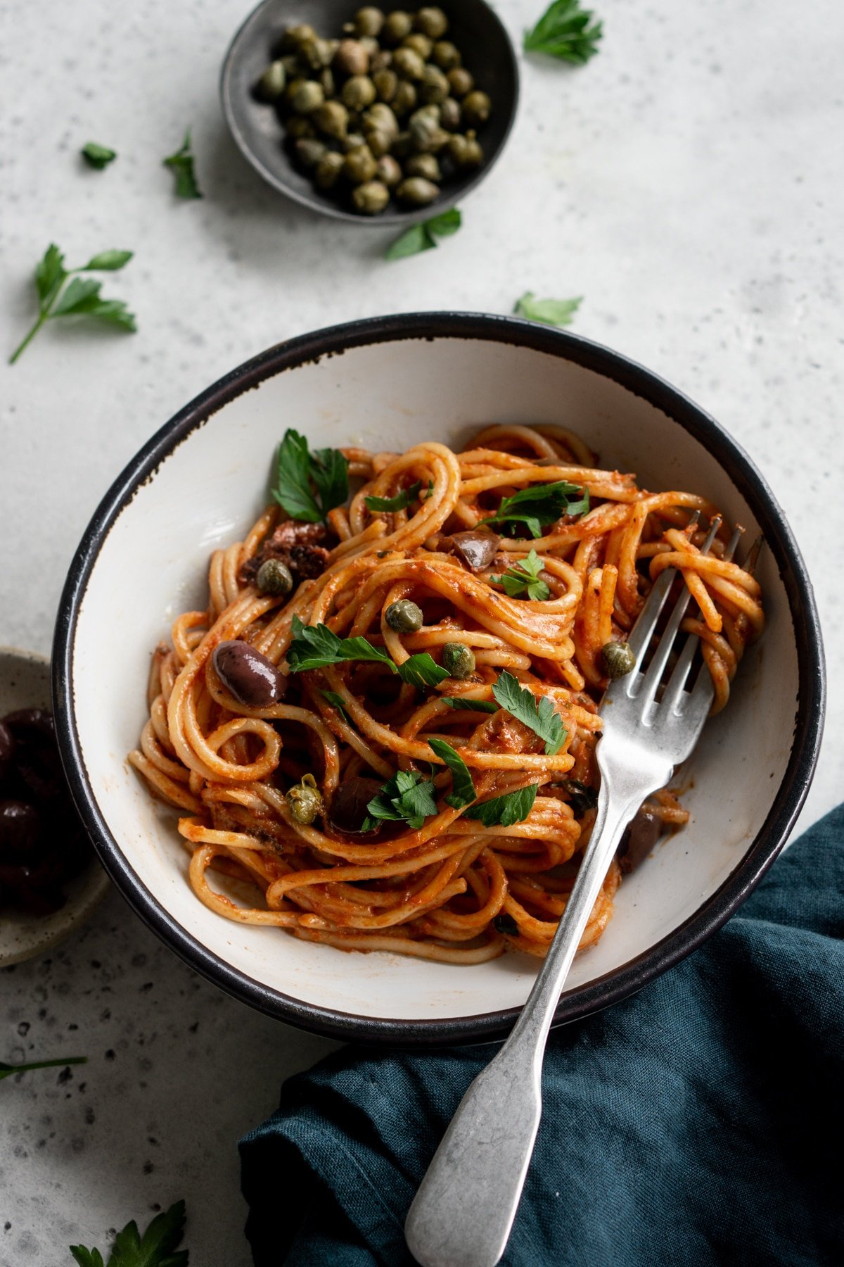 Spaghetti alla puttanesca in a bowl with a small bowl of capers and parsley in the background