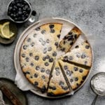 Blueberry ricotta cake in a dish surrounded by a cup of blueberries and lemon wedges