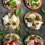 Six mini pizzetta lined up with arugula and basil scattered around