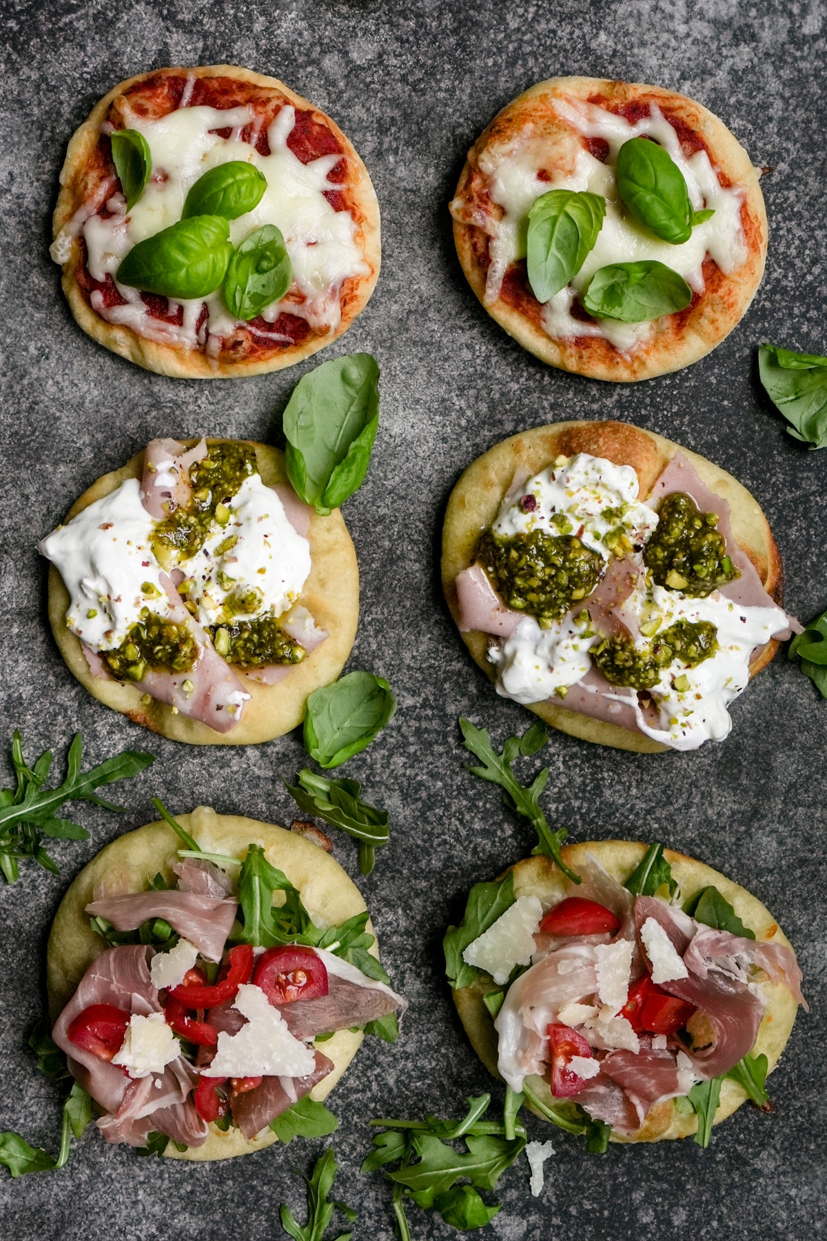 Six mini pizzetta lined up with arugula and basil scattered around