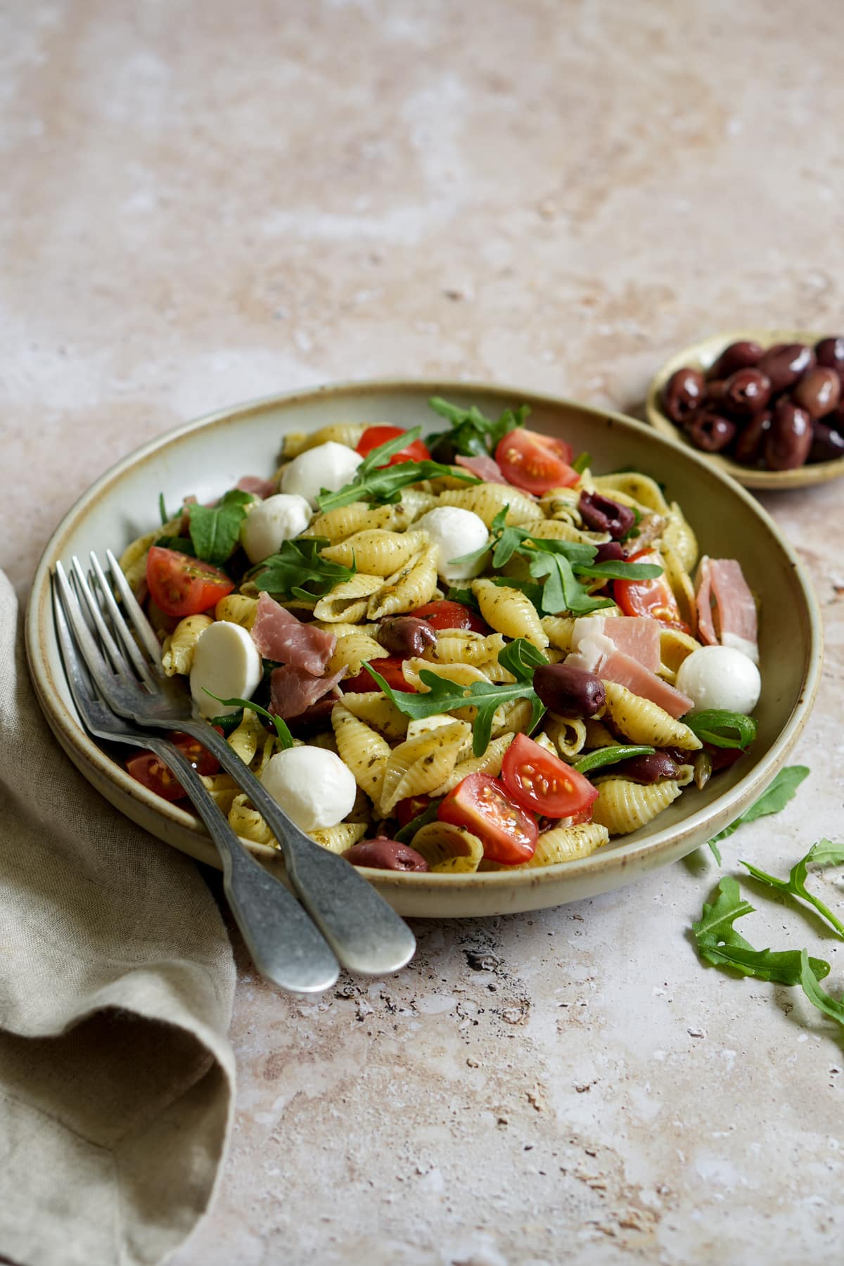 Arugula pasta salad in a bowl topped with prosciutto, mozzarella, tomatoes, and sunflower seeds