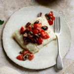 Piece of halibut on a plate topped with tomatoes, olives and capers