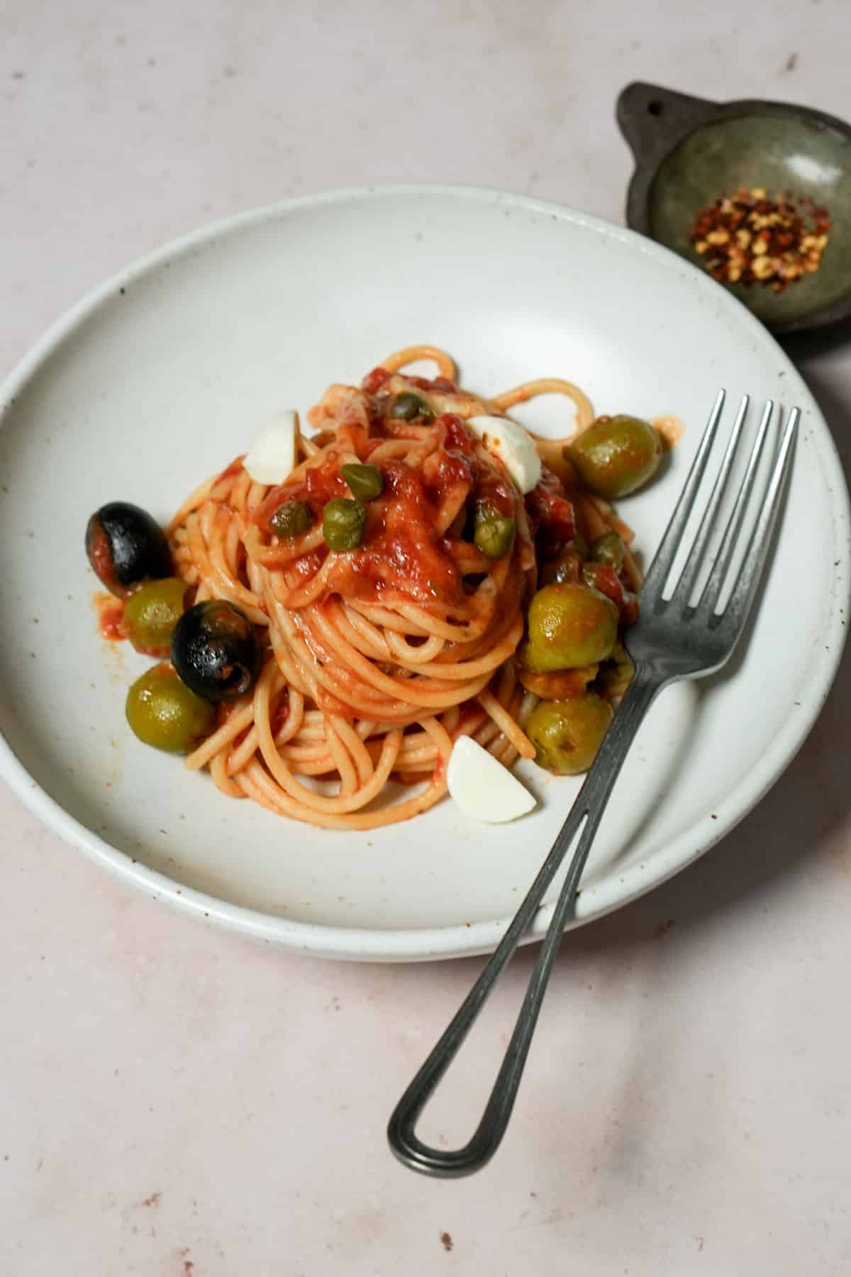 Vesuviana pasta in a dish with a fork, and small bowl of chili flakes in the background