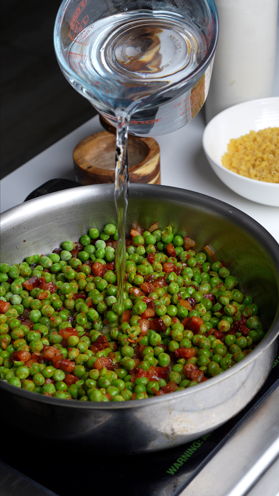 Process photo on how to make pasta e piselli - pancetta, onions and peas in pan with water being poured in
