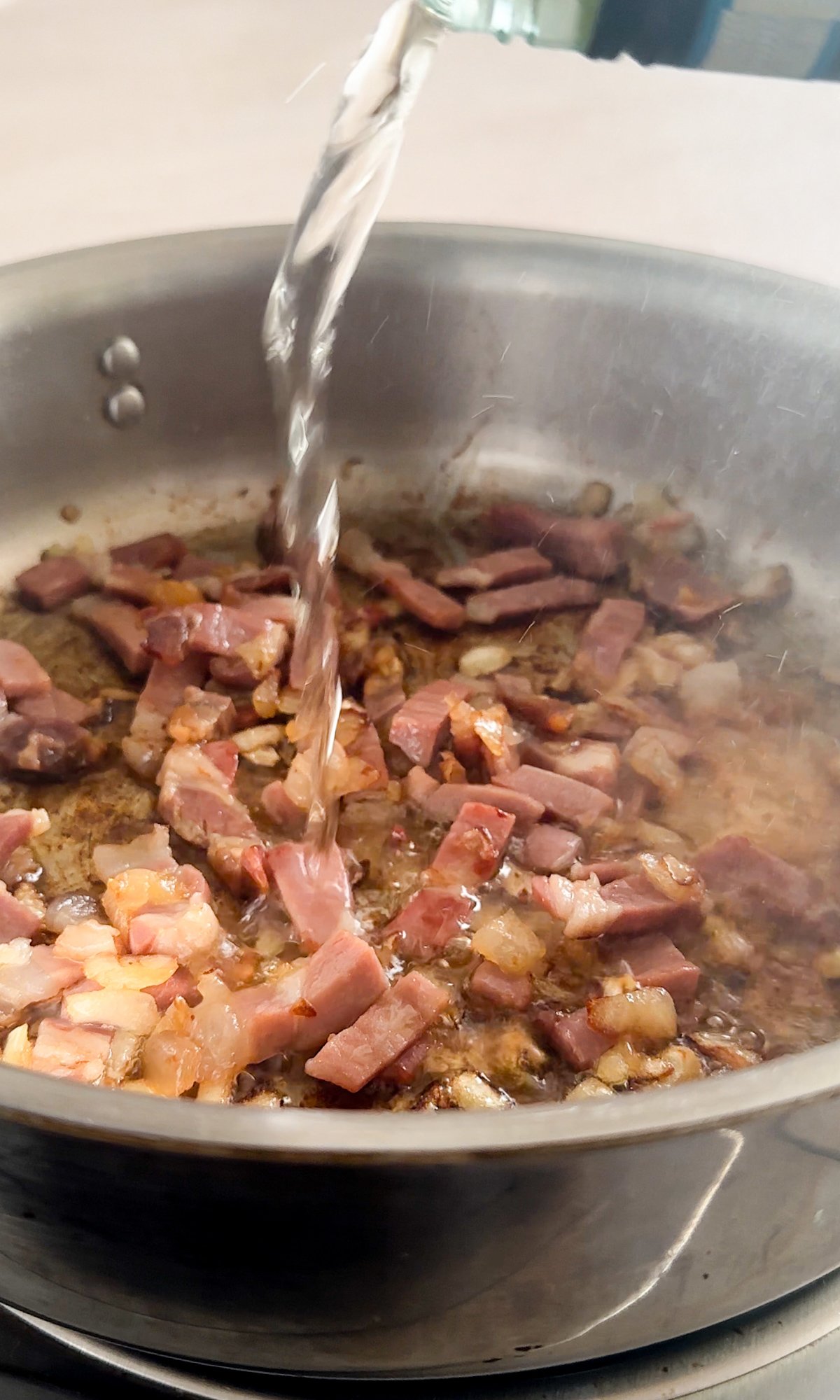 Prosciutto and onions in a pan with white wine being poured in