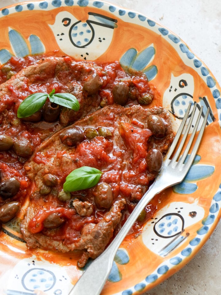 Two thin cut sirloin steaks in a plate topped with tomato sauce and fresh basil with a fork to the side