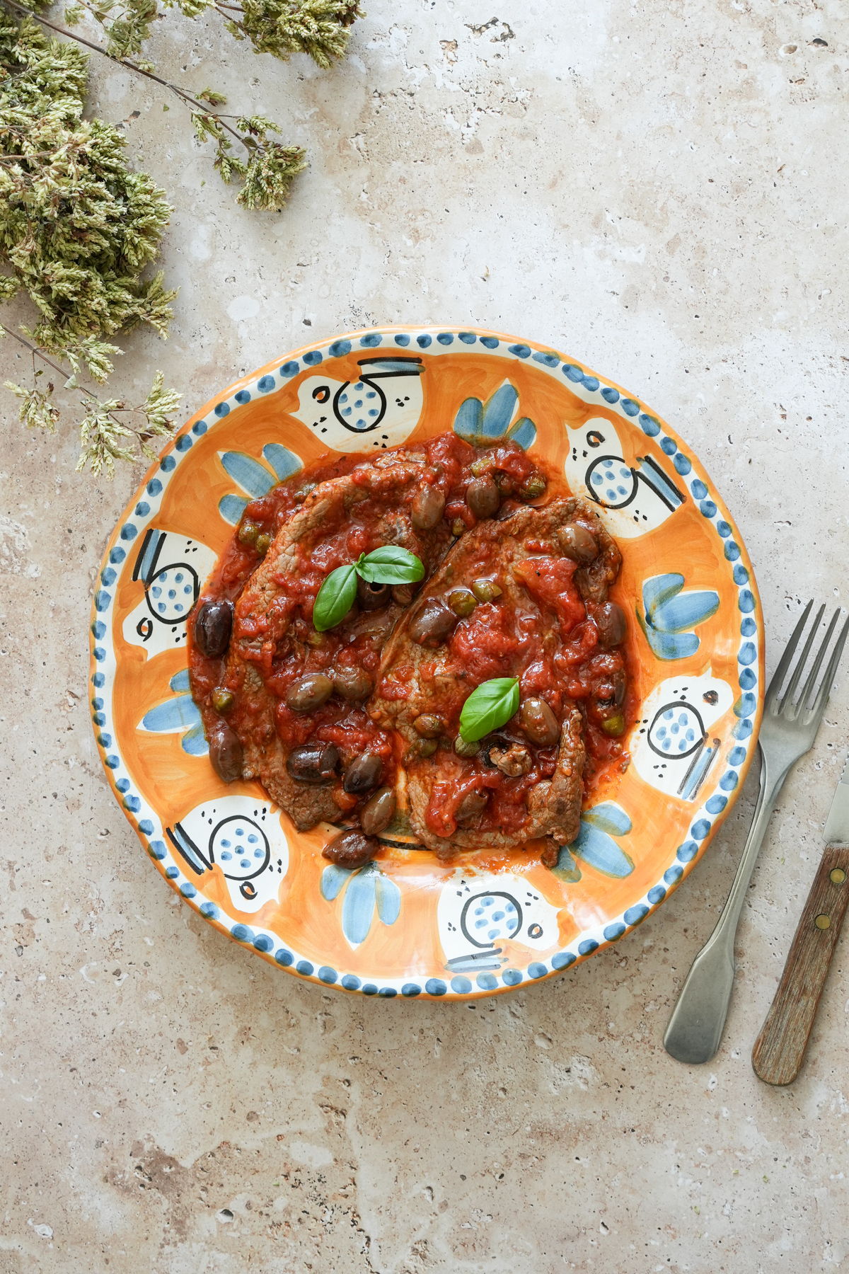 Two thin cut sirloin steaks in a plate topped with tomato sauce, capers, olives and fresh basil