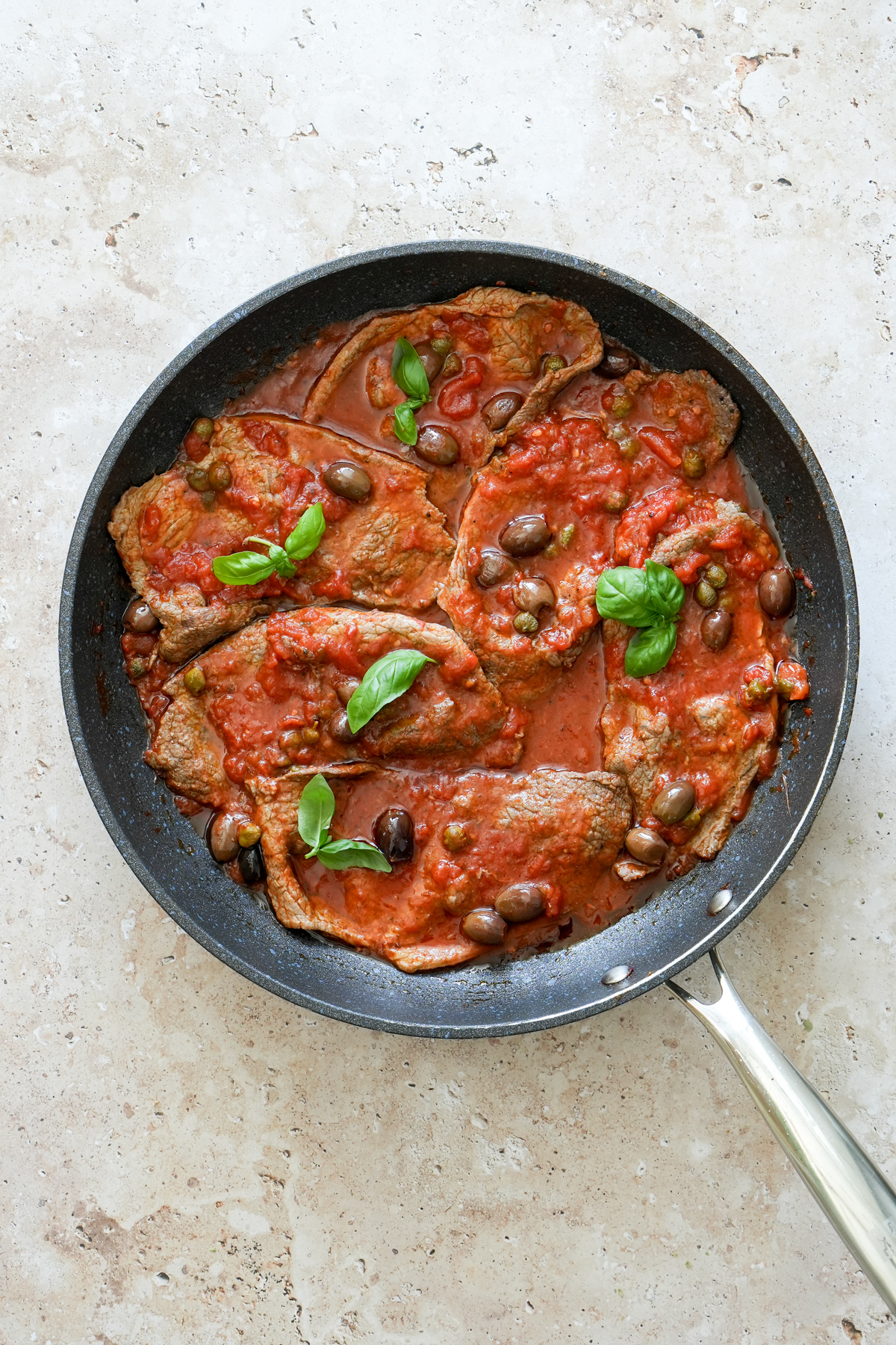 Six thin cut sirloin steaks in a pan with tomato, caper and olive sauce, sprinkled with fresh basil