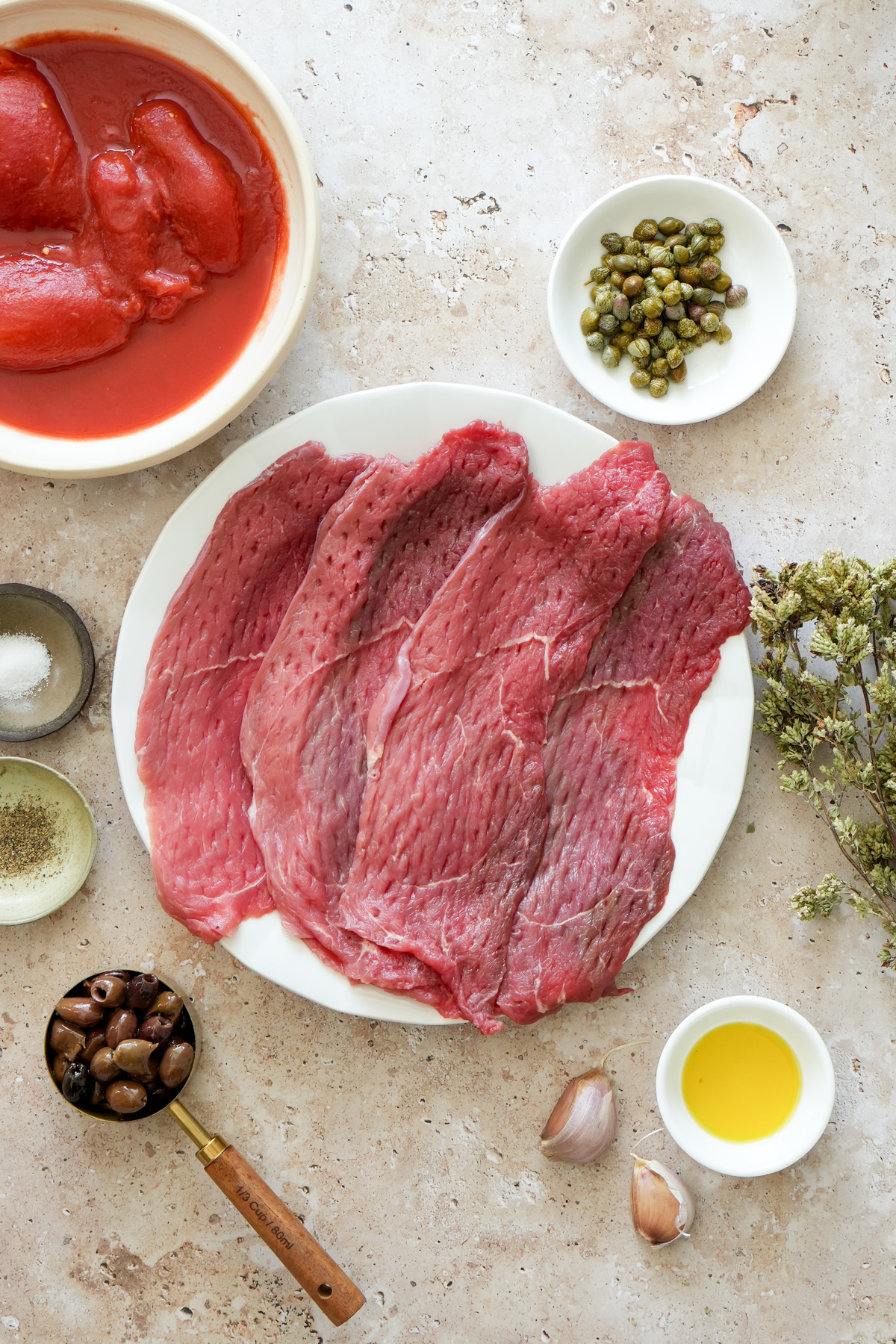 Steak pizzaiola ingredients - thin cut sirloin steaks, capers, dried oregano, olive oil, garlic cloves, olives, salt, pepper, canned tomatoes