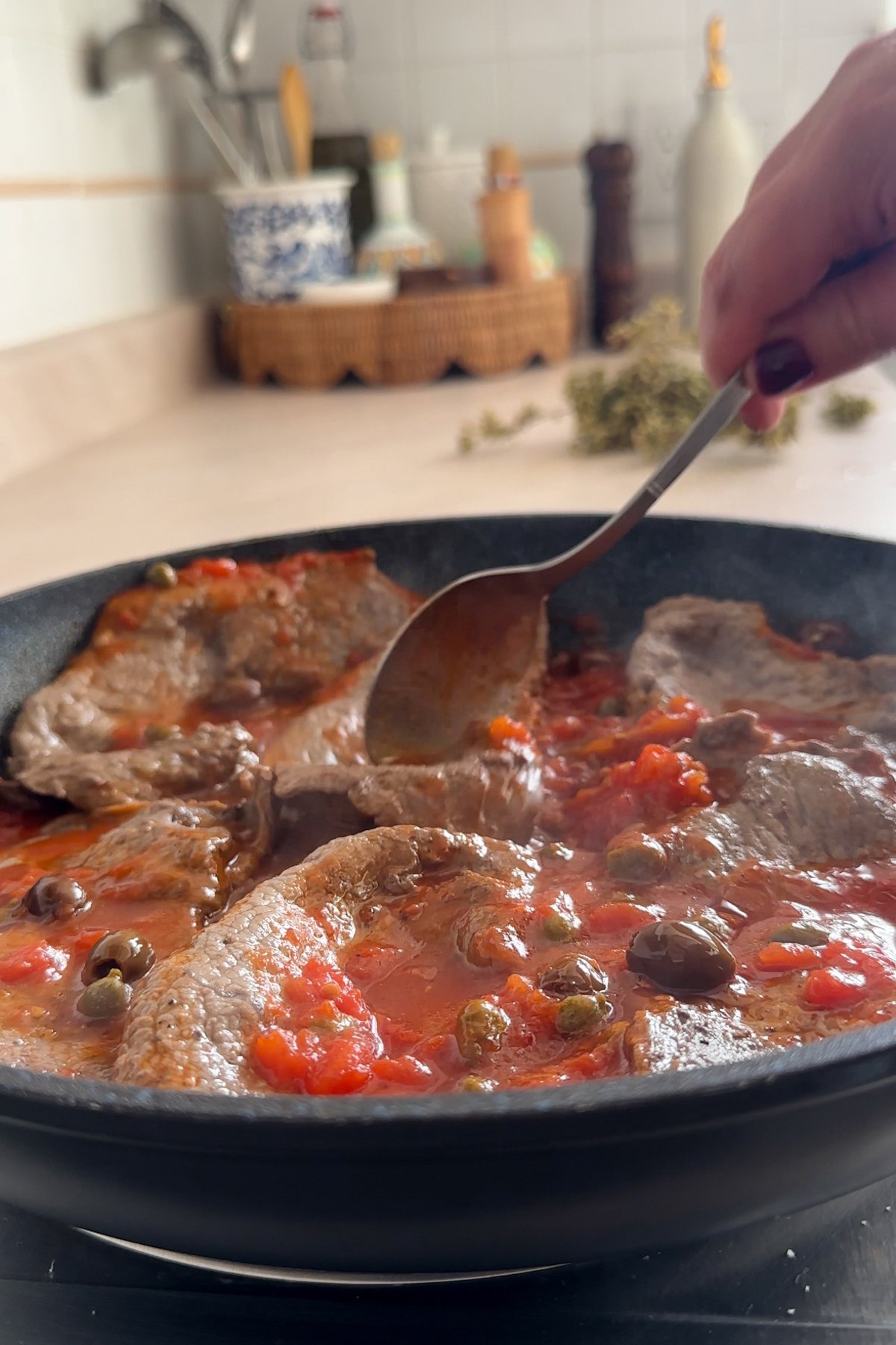 Thin cut sirloin steaks in a pan being topped with tomato sauce, olives and capers