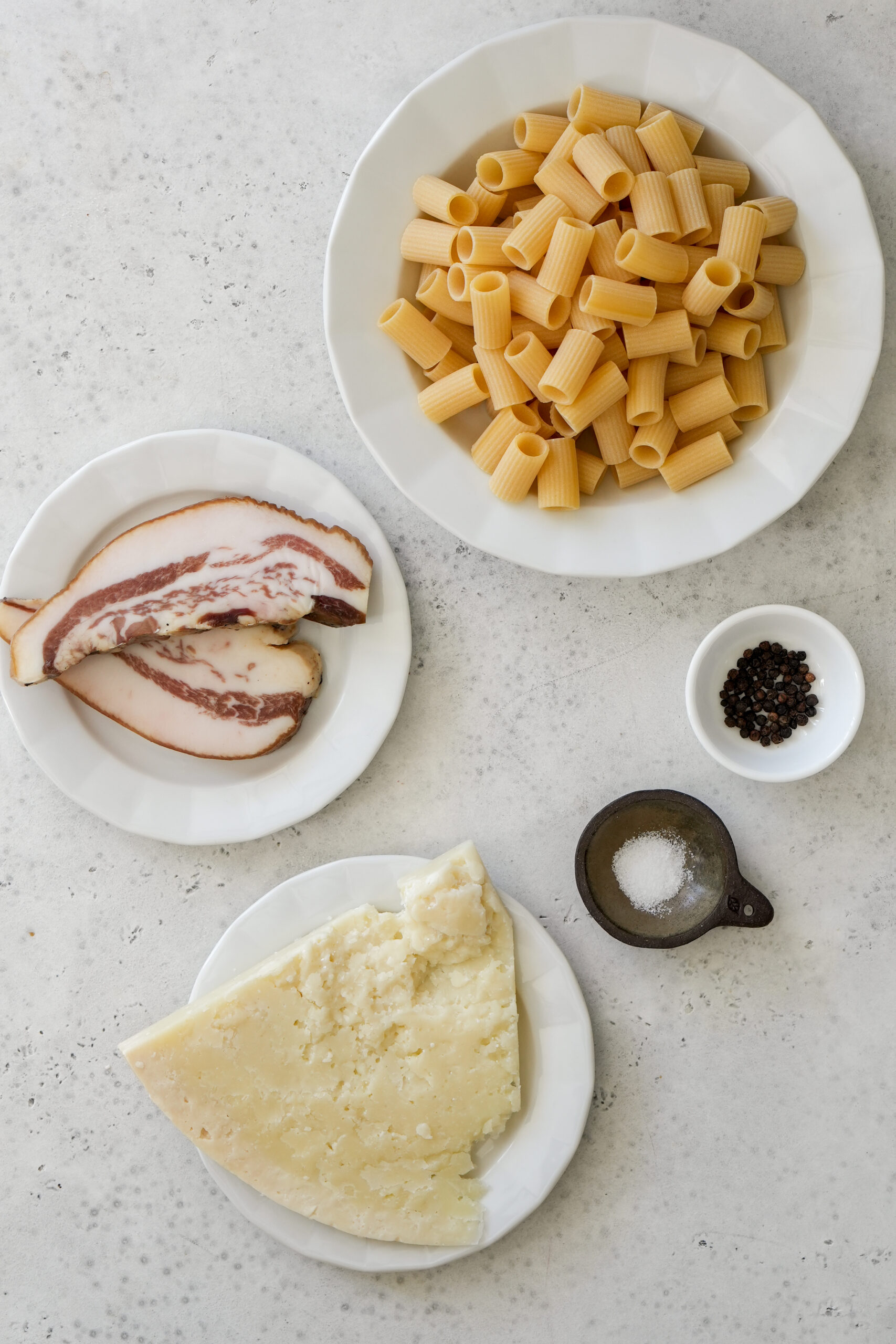 Pasta alla gricia ingredients in individual dishes - mezzi rigatoni, salt, pepper, pecorino and guanciale