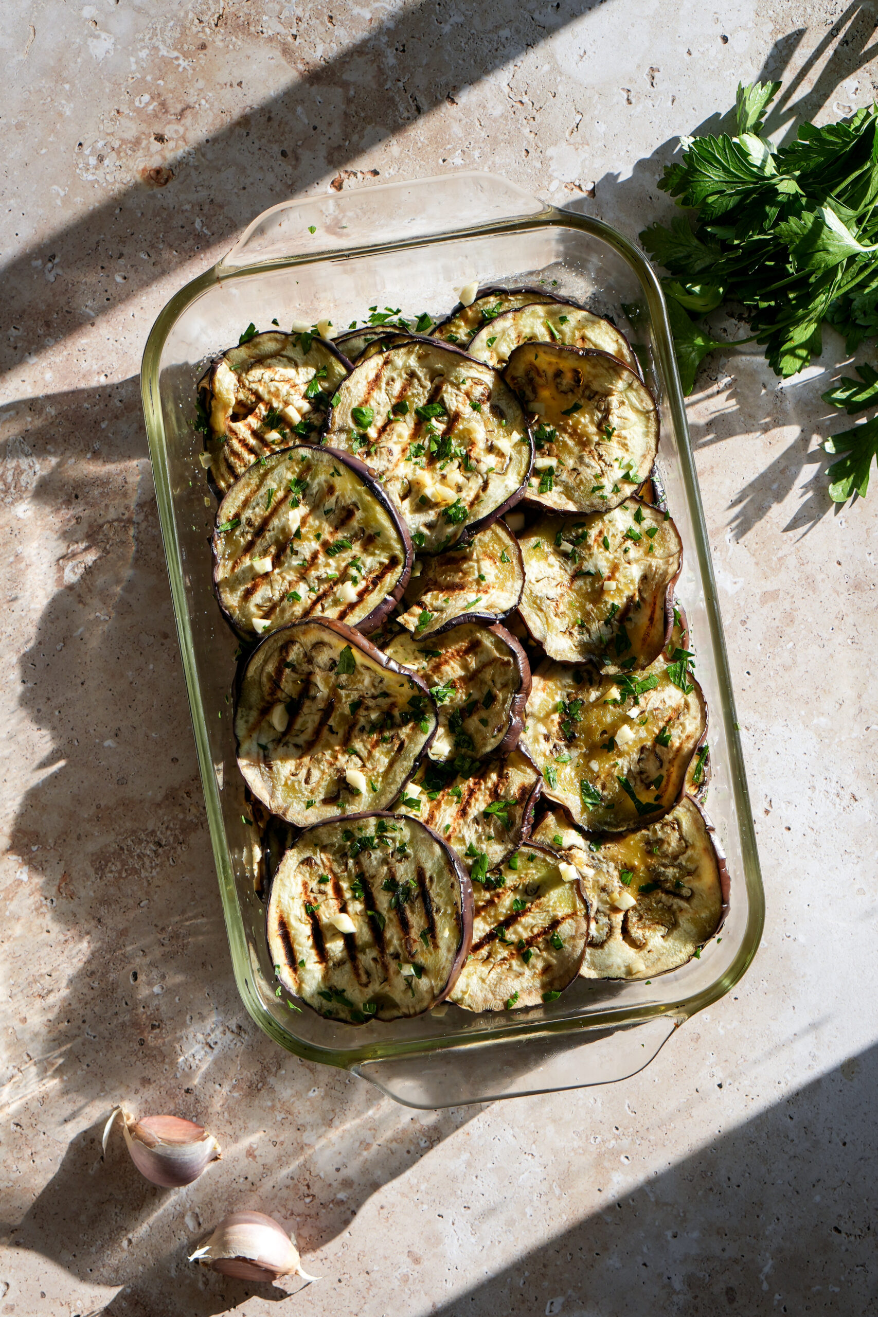 Grilled eggplant rounds in a glass baking dish topped with minced parsley and garlic