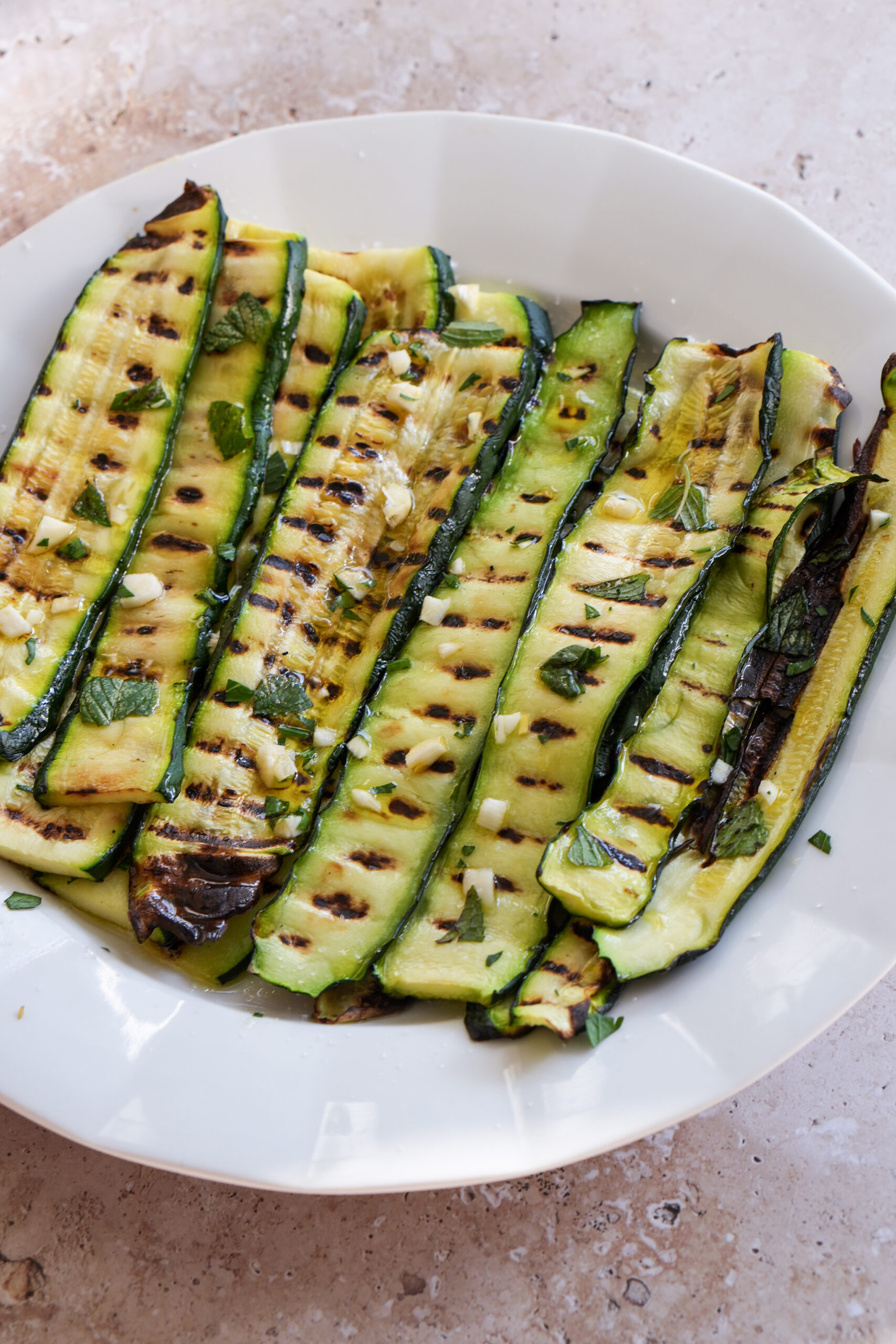 Grilled zucchini in a dish topped with minced garlic, parsley and mint