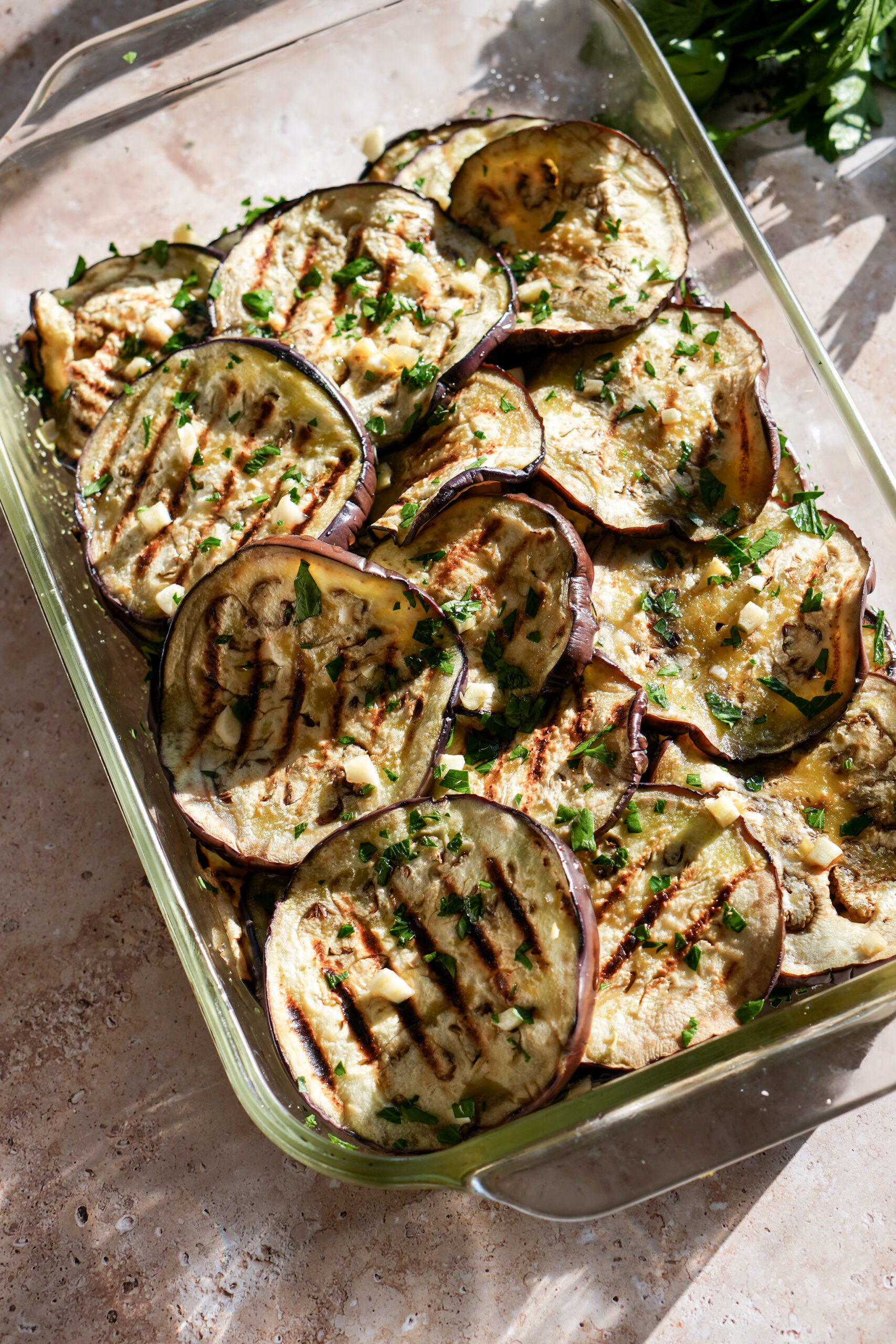 Grilled eggplant in a glass baking dish with fresh parsley in the background
